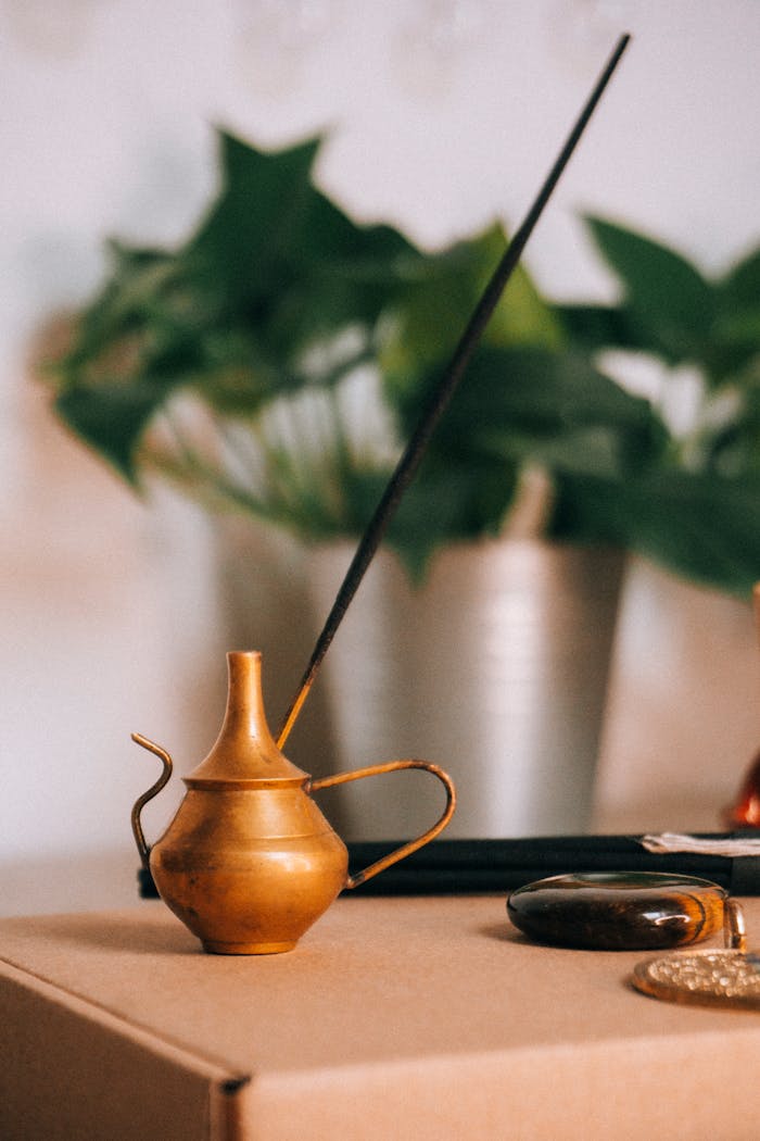 A copper teapot-shaped incense holder with an incense stick, set against a leafy green backdrop indoors.